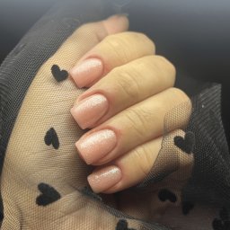 Metamorfoza Salon Urody Maków Mazowiecki Kasia Grabowska Makijaż Rzęsy Paznokcie Brwi - Dłoń z kwadratowymi paznokciami pokrytymi jasnoróżowym lakierem z delikatnym brokatem, trzymająca czarną, przezroczystą tkaninę w serduszka.