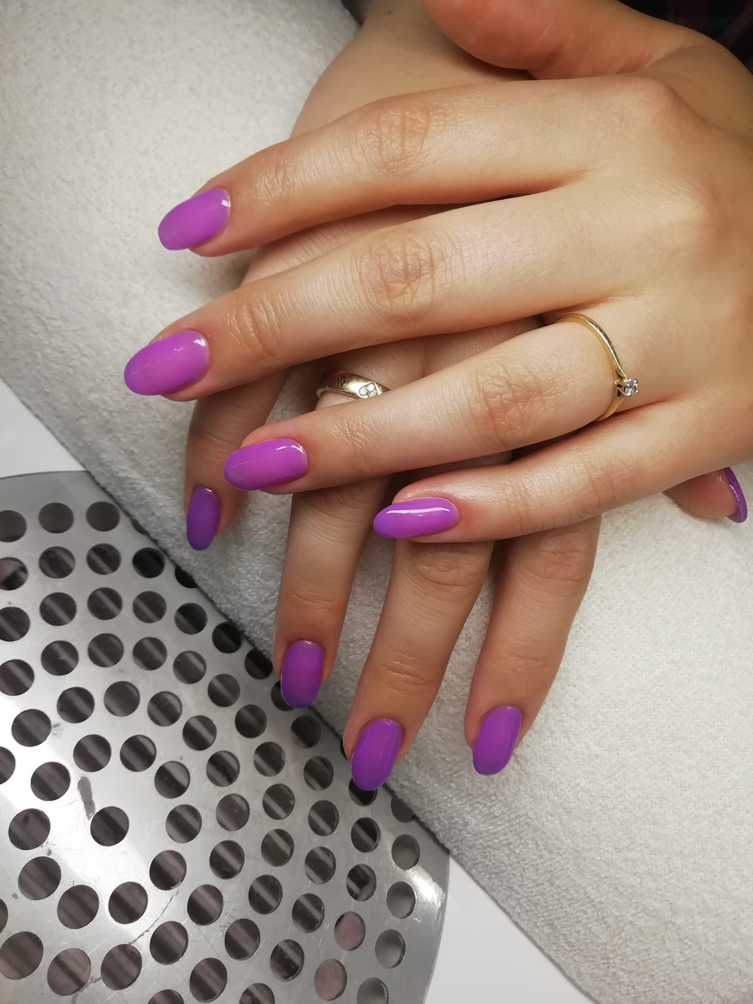 Dłonie kobiety z owalnymi paznokciami pomalowanymi na jasny fiolet, na palcu złoty pierścionek z pojedynczym kamieniem, tło stanowi biały ręcznik i metalowa kratka wentylacyjna.