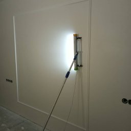 Taurus Denys Sushniak - Szlifowanie ściany szlifierką z oświetleniem punktowym, widoczne wykończenia dekoracyjne i gniazdka elektryczne. Prace wykończeniowe w jasnym pomieszczeniu.