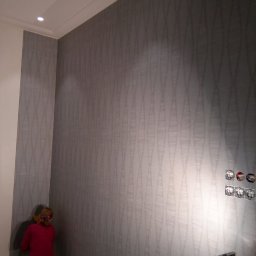 Taurus Denys Sushniak - Tapetowanie ściany z wzorem w odcieniach szarości. Kobieta w trakcie pracy, widoczne reflektory i gniazdka elektryczne. Wykończenie wnętrza w trakcie realizacji.