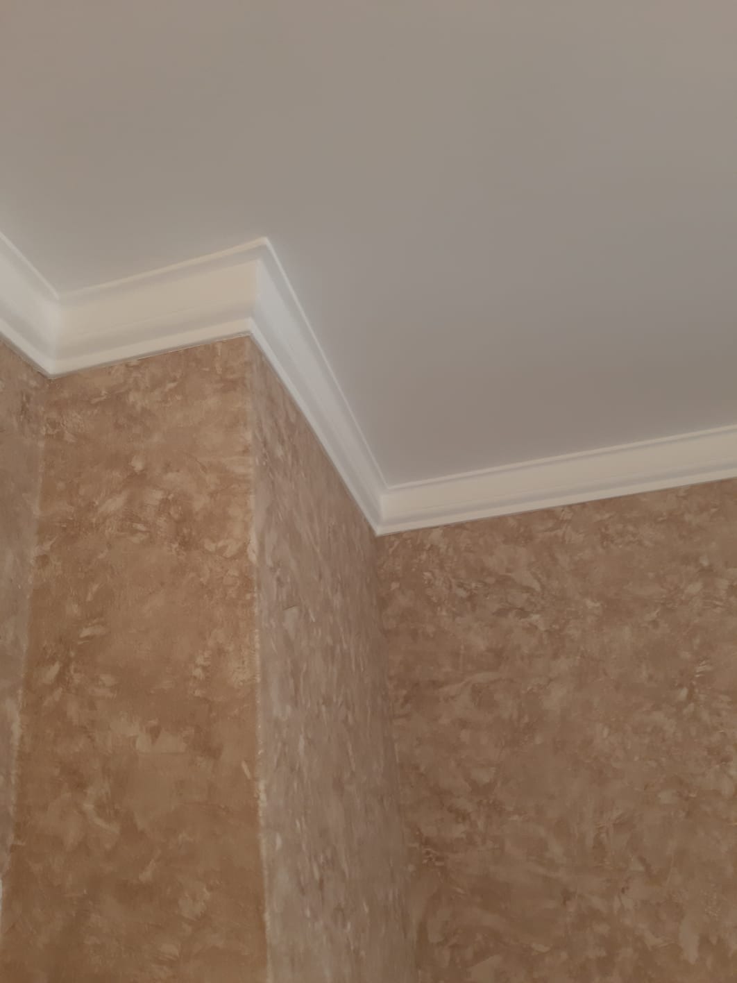 Wykończony narożnik pokoju z dekoracyjną, beżową ścianą i białym, profilowanym wykończeniem sufitu. Detal wykonania.