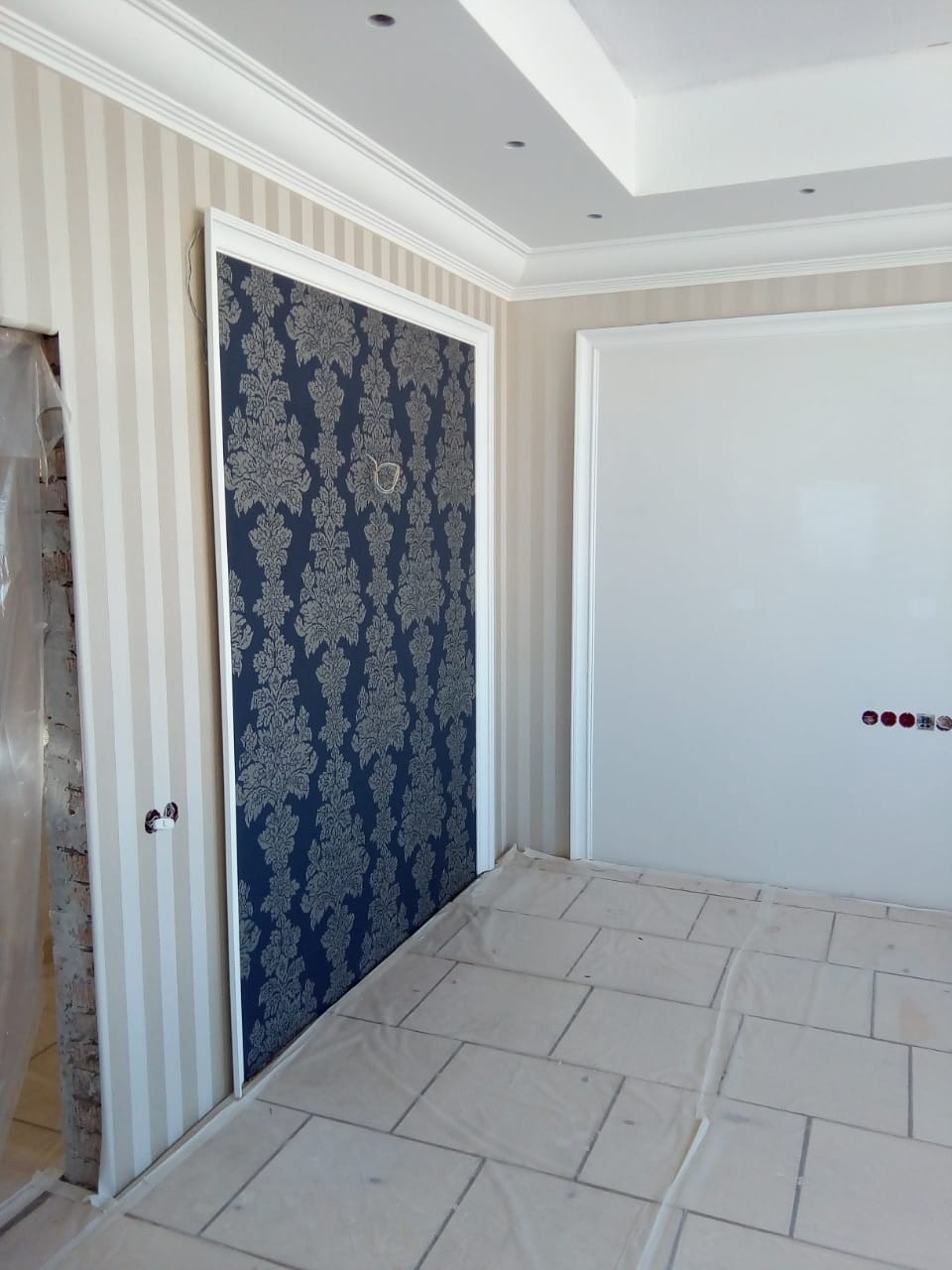 Pokój po remoncie: wzorzysta tapeta na ścianie, listwy przypodłogowe, zabezpieczona podłoga. Widoczne gniazdka elektryczne i otwór w ścianie.