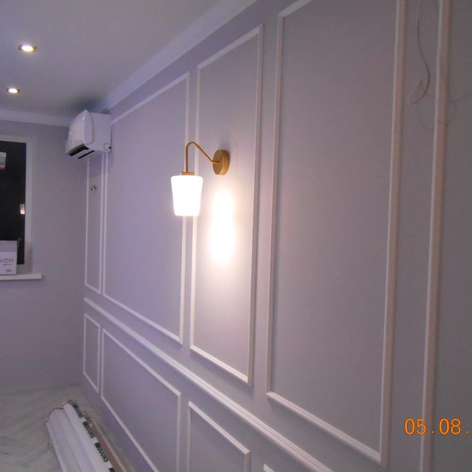 Ściana w odcieniach szarości z dekoracyjnymi listwami i złotym kinkietem. Widoczny klimatyzator i fragment podłogi. Eleganckie wykończenie wnętrza.