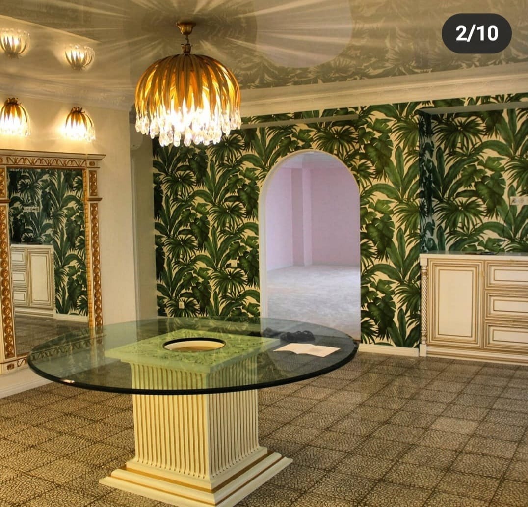 Eklektyczne wnętrze: okrągły stół z blatem szklanym i zielonym marmurem, tropikalna tapeta z motywem liści, złoty żyrandol i szafka w stylu vintage.