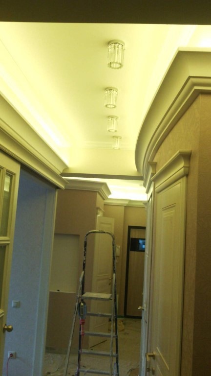 Korytarz w trakcie wykańczania: widoczny sufit z oświetleniem punktowym i dekoracyjnymi listwami, ściany w odcieniach beżu, drabina malarska.