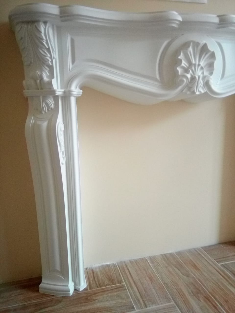 Ozdobny, biały portal kominkowy z dekoracyjnymi rzeźbieniami, zamontowany we Wrocławiu. Klasyczny styl, kremowe ściany i drewnopodobna podłoga.