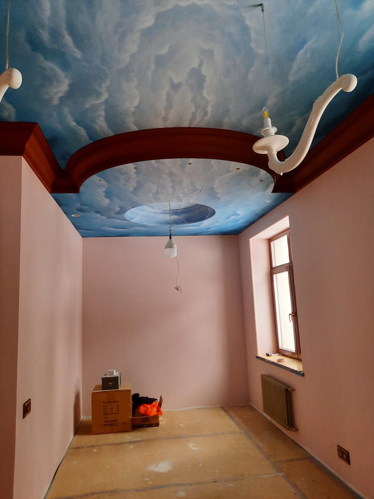 Pokój po malowaniu ścian na różowo i artystycznym malowaniu sufitu w błękitne chmury. Widoczne dekoracyjne elementy oświetleniowe i wykończeniowe.
