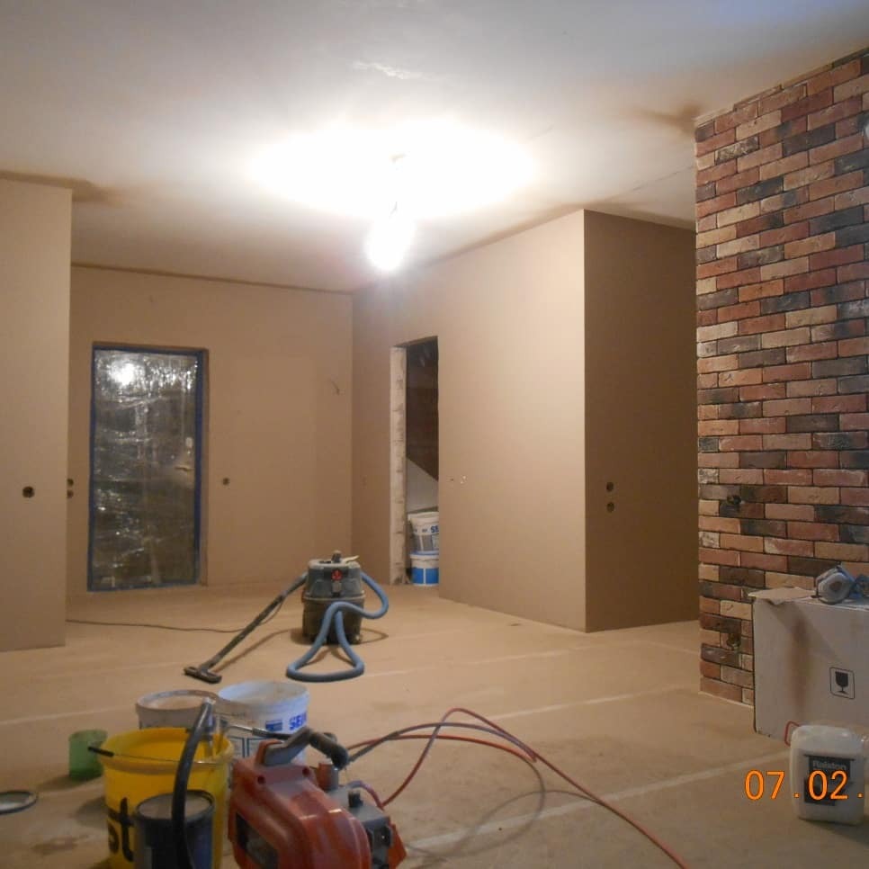 Wnętrze w trakcie prac wykończeniowych: ściany po szpachlowaniu, przygotowane do malowania. Widoczne narzędzia i materiały budowlane, fragment ściany z cegły dekoracyjnej.