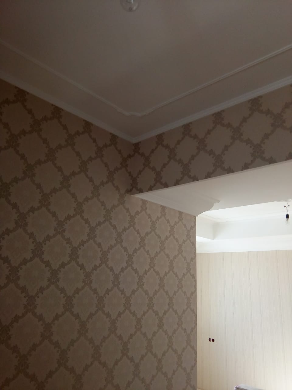 Wnętrze po remoncie: ściana w beżowe wzory z dekoracyjną listwą pod sufitem. Widoczny fragment drugiego pomieszczenia z jasnymi ścianami i wystającymi kablami.