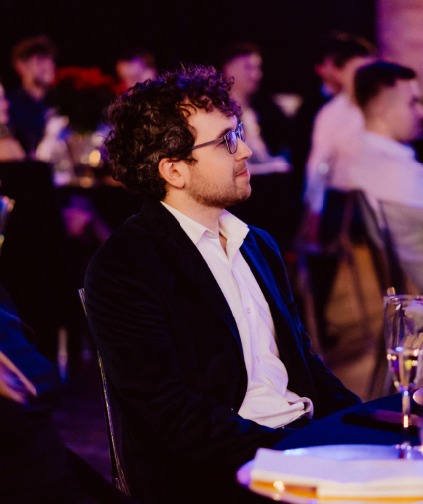 Młody mężczyzna w okularach i czarnej marynarce, z kręconymi włosami, siedzi na krześle w pomieszczeniu z przyciemnionym oświetleniem, patrząc w prawo. W tle widoczni inni ludzie.