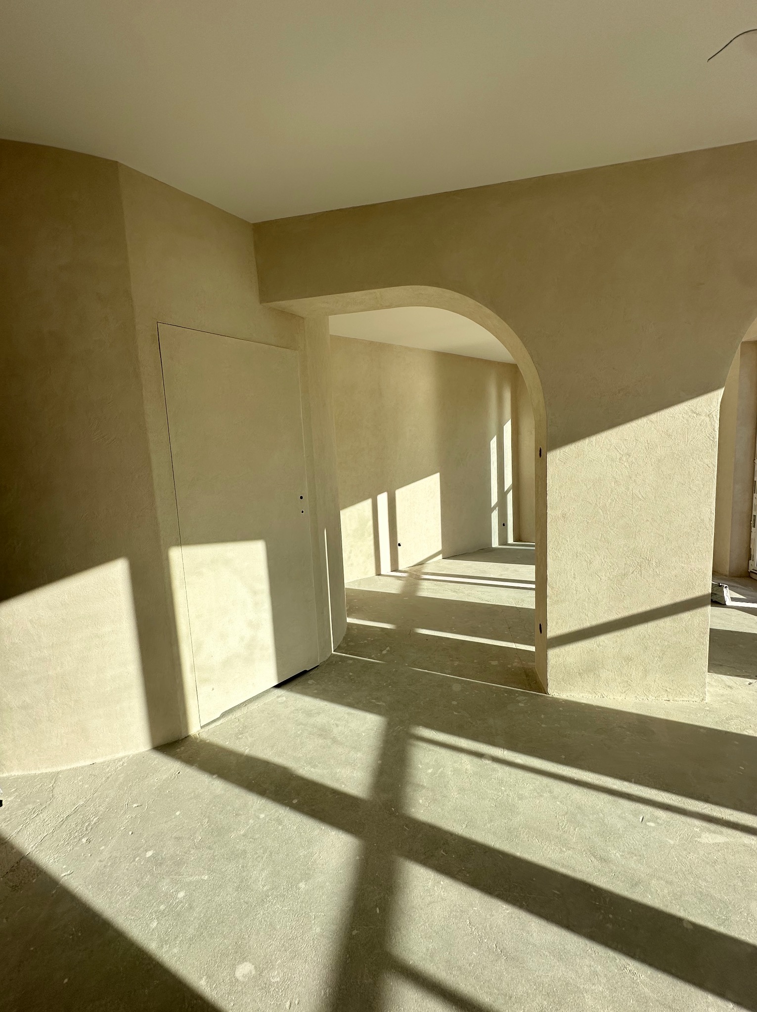 Wnętrze w trakcie prac wykończeniowych, świeżo położone jasne gładzie, widoczne promienie słoneczne wpadające przez okno, surowy beton na podłodze, charakterystyczne łukowe przejście.
