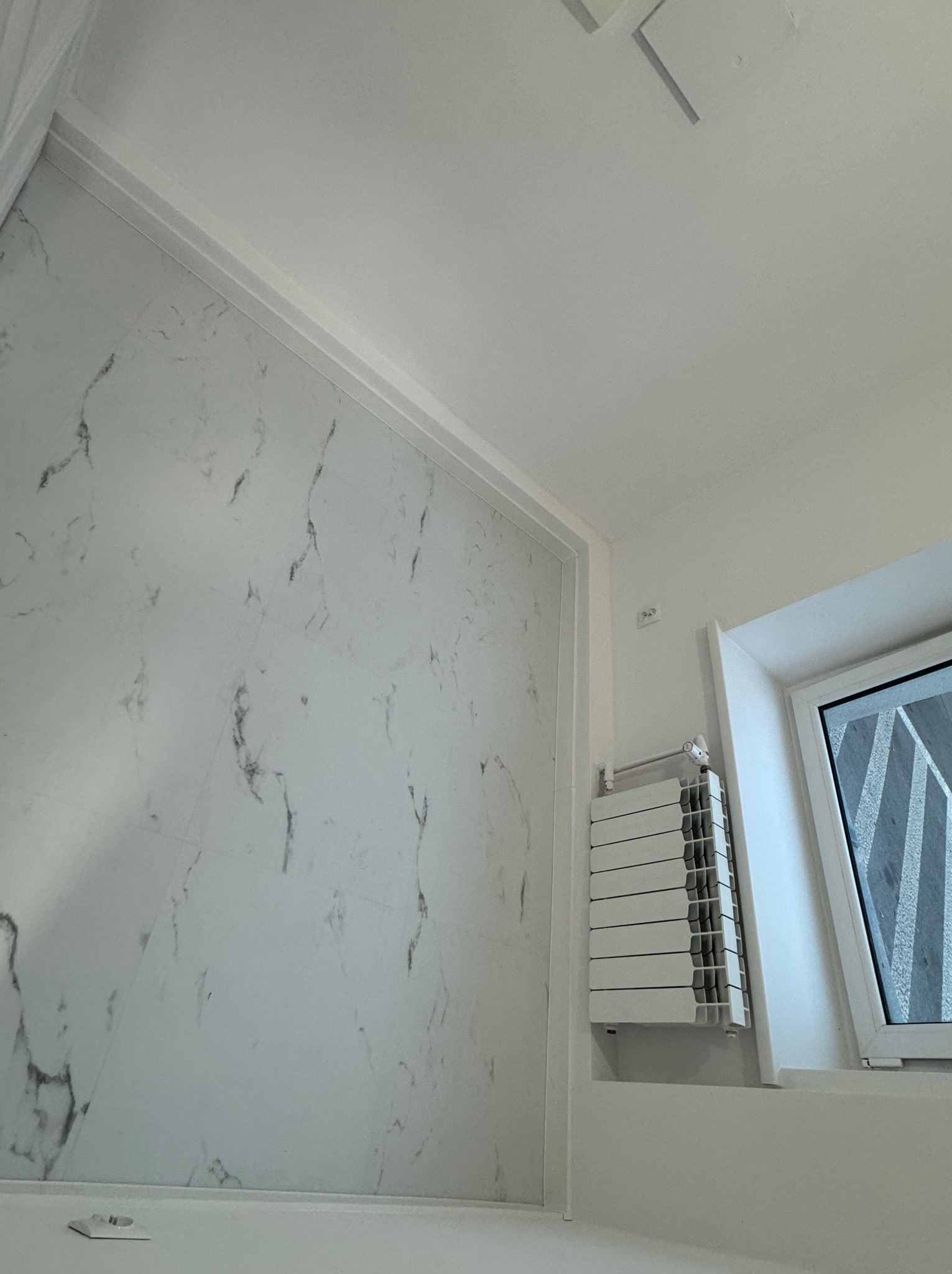 Wykończona łazienka z marmurową ścianą, grzejnikiem żeberkowym pod oknem i minimalistycznym oświetleniem sufitowym.