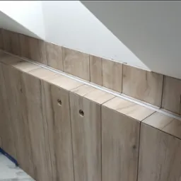 Zabudowa pod skosem z drewnopodobnych paneli z widocznymi otworami na uchwyty, wykończona aluminiową listwą, na tle białej ściany poddasza.