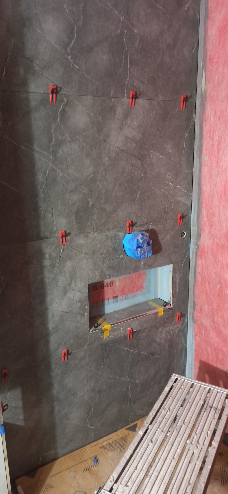 Montaż płytek imitujących marmur na ścianie z wyciętą wnęką, zabezpieczone niebieską folią, użyto klipsów poziomujących i krzyżyków dystansowych, widoczny fragment rusztowania.