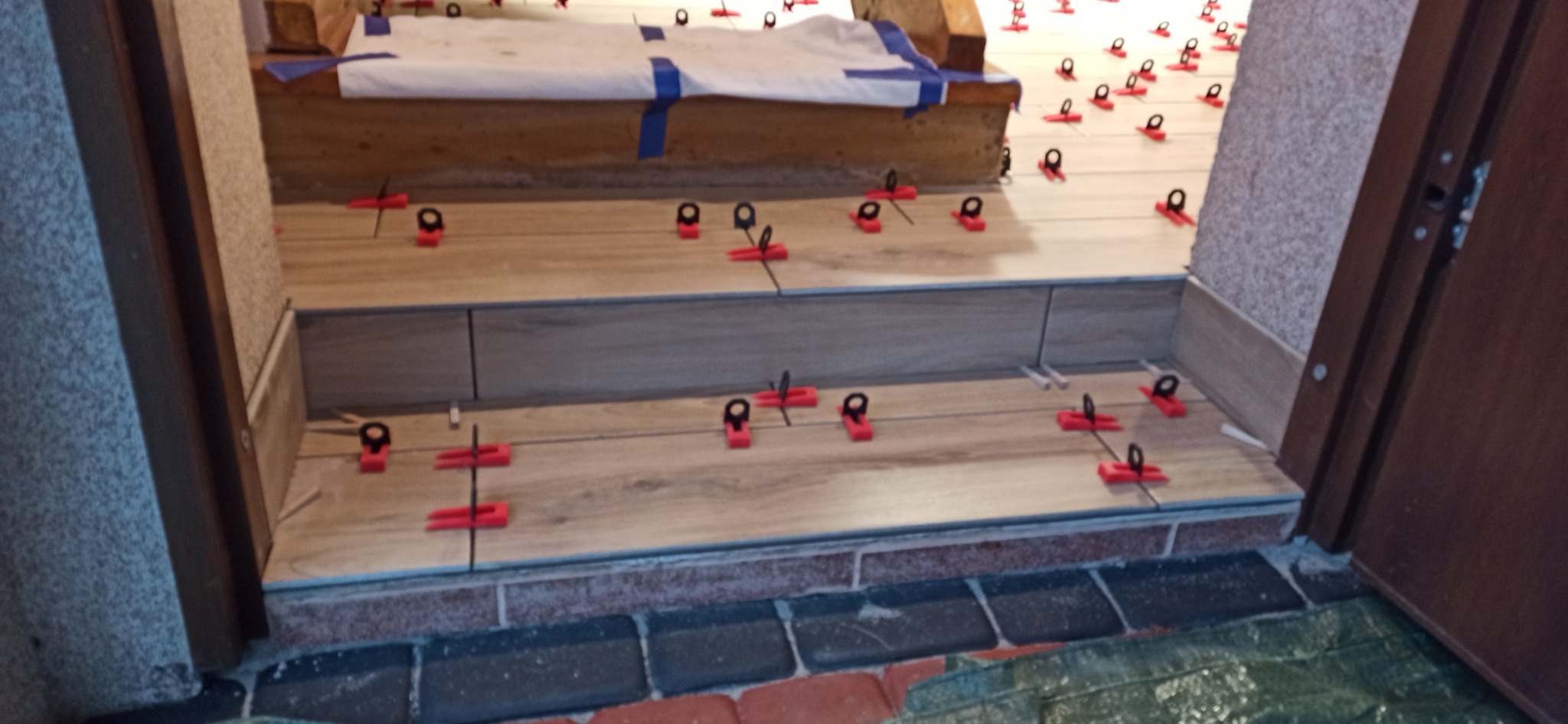 Montaż płytek podłogowych imitujących drewno na schodach wejściowych, z użyciem czerwonych klipsów poziomujących i białej zaprawy, widoczna ceglana podmurówka i fragment drzwi.