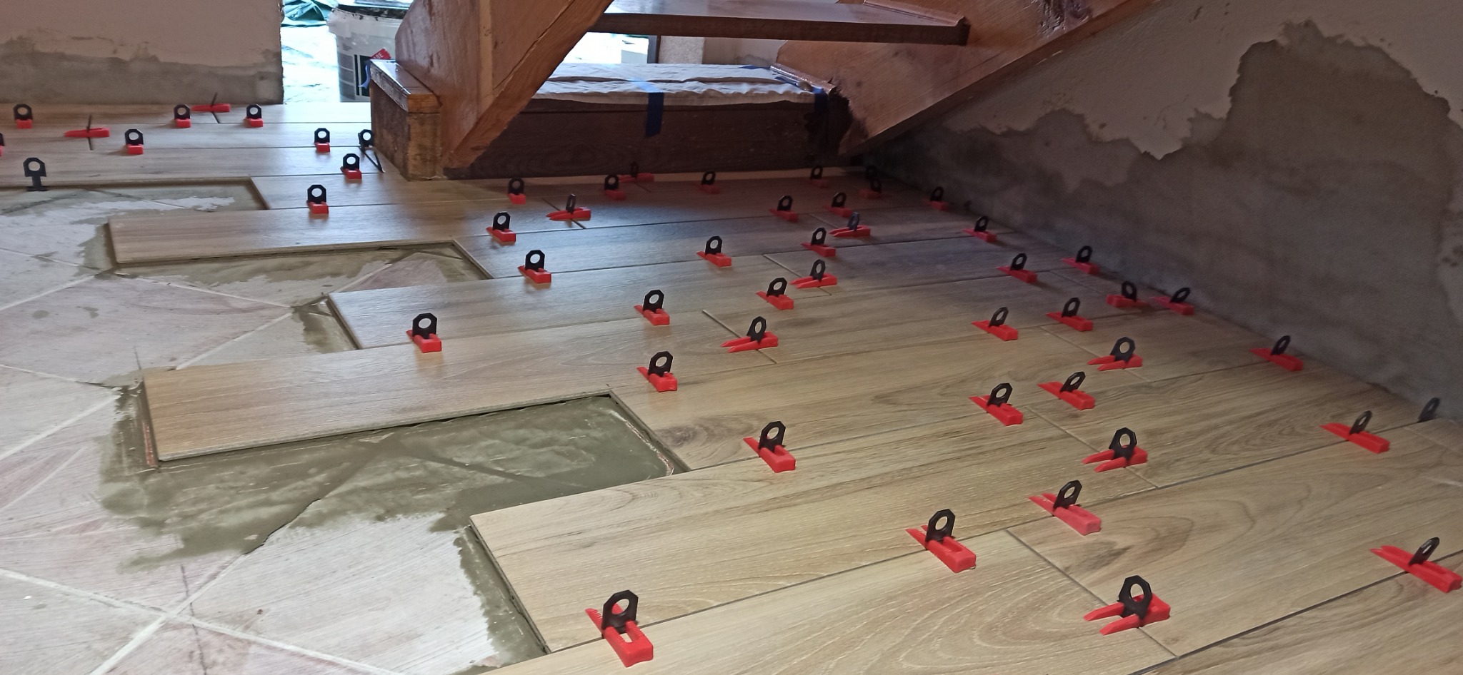 Układanie podłogi z płytek imitujących drewno, z użyciem czerwonych klipsów poziomujących, widoczne pozostałości zaprawy i fragment schodów.