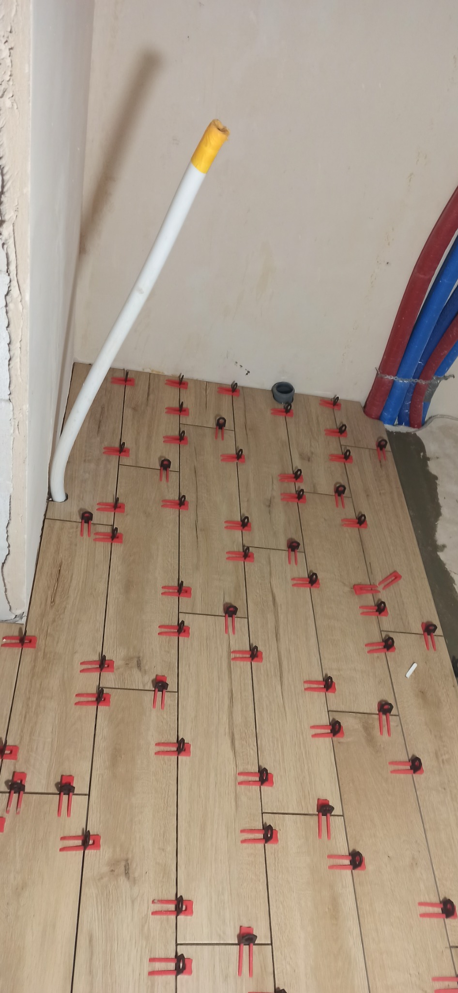 Układanie płytek podłogowych imitujących drewno z użyciem czerwonych klipsów poziomujących, widoczne rury instalacyjne i fragment ściany w trakcie prac wykończeniowych.