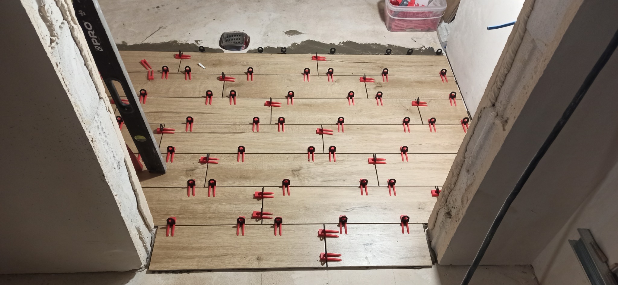 Układanie podłogi z płytek imitujących drewno z użyciem klipsów poziomujących; widoczny poziomica przy ścianie i odpływ liniowy.