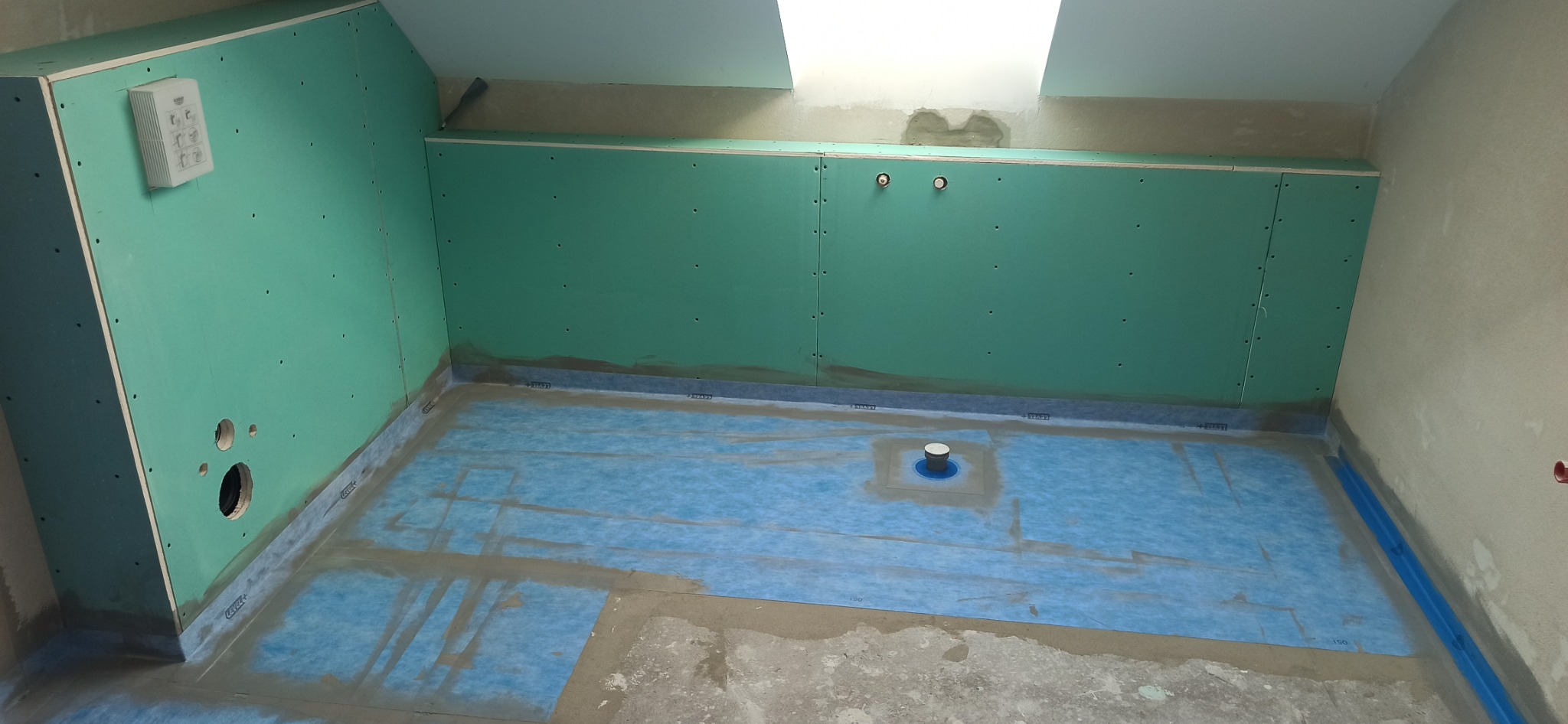 Przygotowanie łazienki w poddaszu do położenia płytek: ściany wyłożone płytami gipsowo-kartonowymi w kolorze zielonym, na podłodze niebieska membrana wodoszczelna z odpływem liniowym.