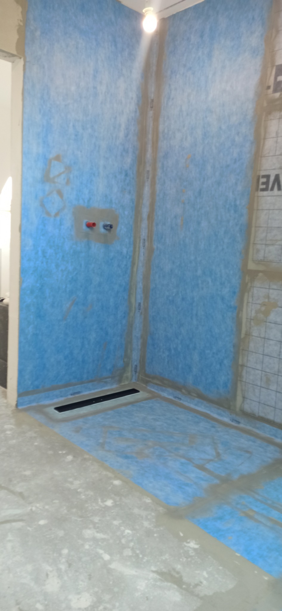 Przygotowanie do hydroizolacji kabiny prysznicowej: ściany i podłoga pokryte niebieską membraną w płynie, widoczne odpływ liniowy i wyprowadzenia instalacji wodnej.