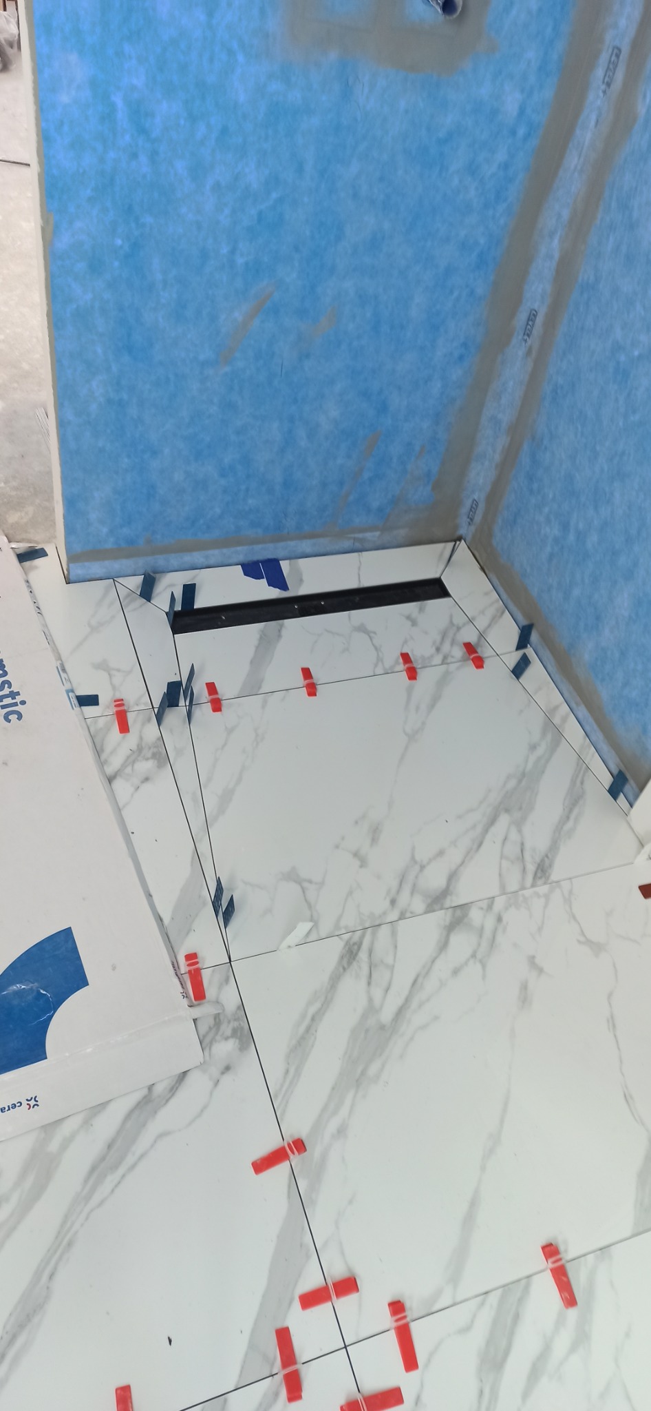 Montaż płytek imitujących marmur w kabinie prysznicowej z odpływem liniowym, widoczne krzyżyki dystansowe i niebieska hydroizolacja na ścianach.