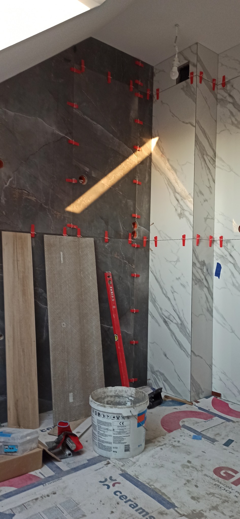 Układanie płytek imitujących marmur i kamień w łazience, widoczne krzyżyki dystansowe, poziomica, wiadro z klejem i nieułożone płytki