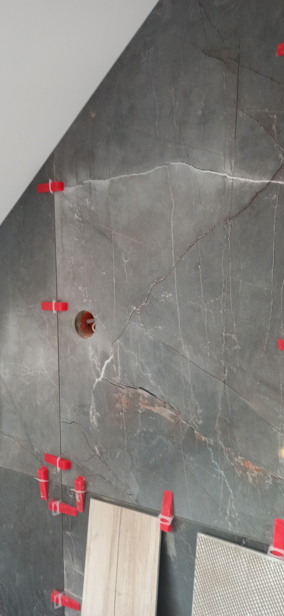 Układanie szarych płytek imitujących marmur na skośnej ścianie, widoczne czerwone kliny poziomujące i otwór na rurę.