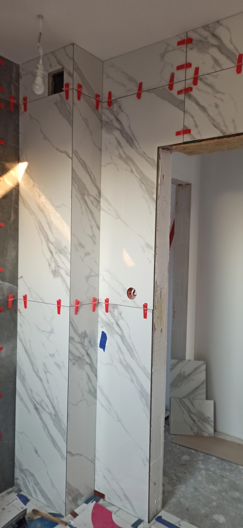 Ściana w trakcie wykańczania białymi płytkami imitującymi marmur, z czerwonymi klipsami poziomującymi i niebieską taśmą, widoczne niedokończone gniazdko elektryczne i surowe wykończenie futryny drzwi.