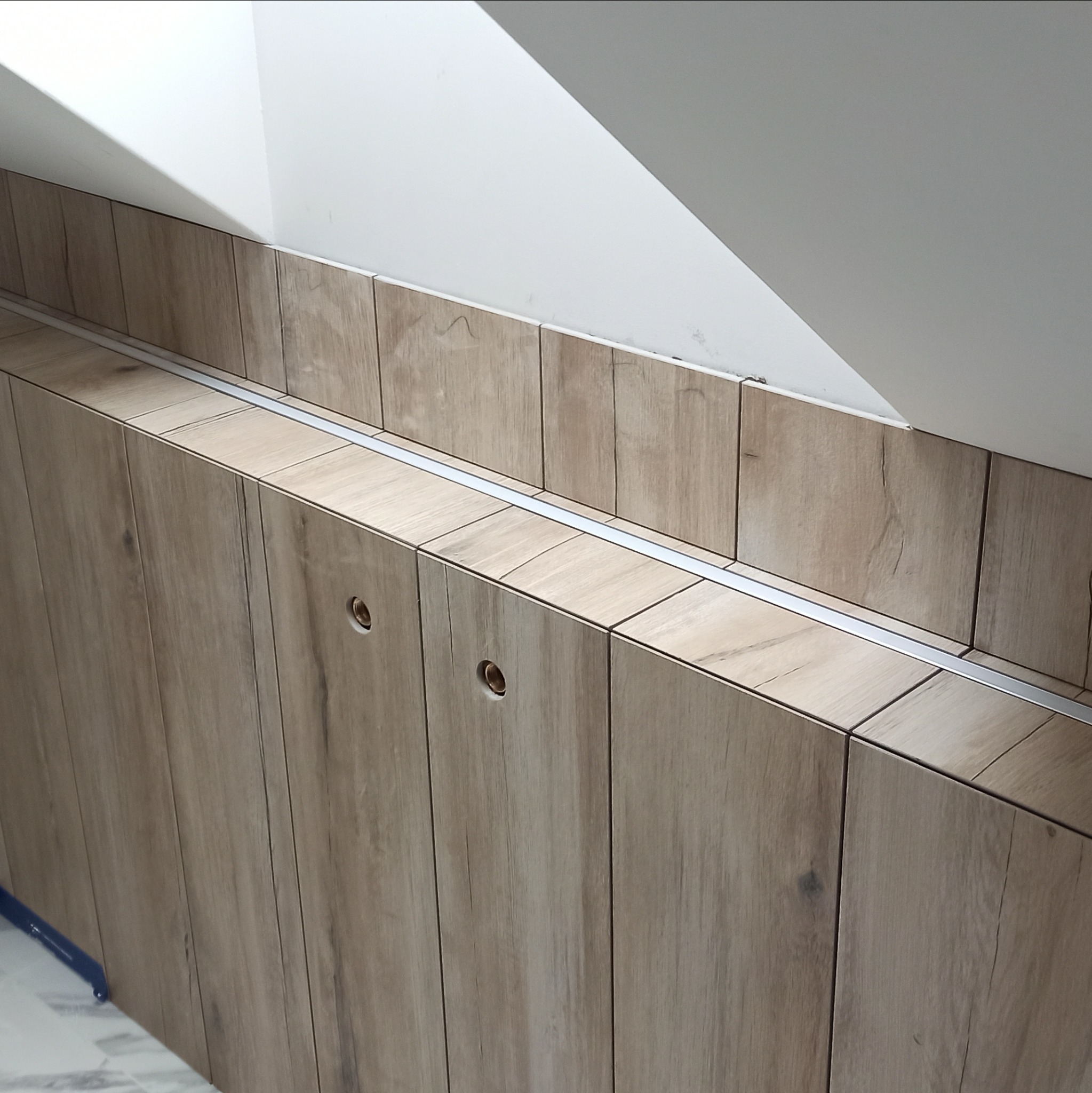 Zabudowa pod skosem z drewnopodobnych paneli z widocznymi otworami na uchwyty, wykończona aluminiową listwą, na tle białej ściany poddasza.