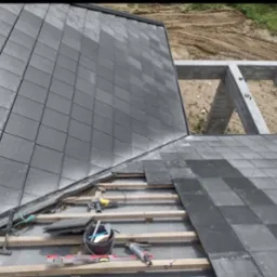 Montaż ciemnoszarej dachówki na drewnianej konstrukcji dachu, widoczne narzędzia pracy i fragment betonowej konstrukcji domu w tle.