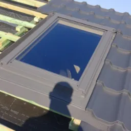 Montaż okna dachowego na nowym dachu z ciemnej blachy falistej, widoczna więźba i membrana dachowa, cień osoby fotografującej na dachu, błękitne niebo.