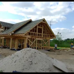 Wznoszenie drewnianej konstrukcji domu jednorodzinnego z częściowo ułożonym pokryciem dachowym z dachówki ceramicznej, widoczny szkielet więźby dachowej i ściany, na pierwszym planie pryzma piasku...