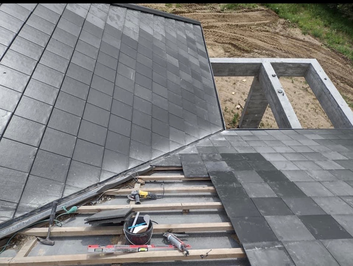 Montaż ciemnoszarej dachówki na drewnianej konstrukcji dachu, widoczne narzędzia pracy i fragment betonowej konstrukcji domu w tle.