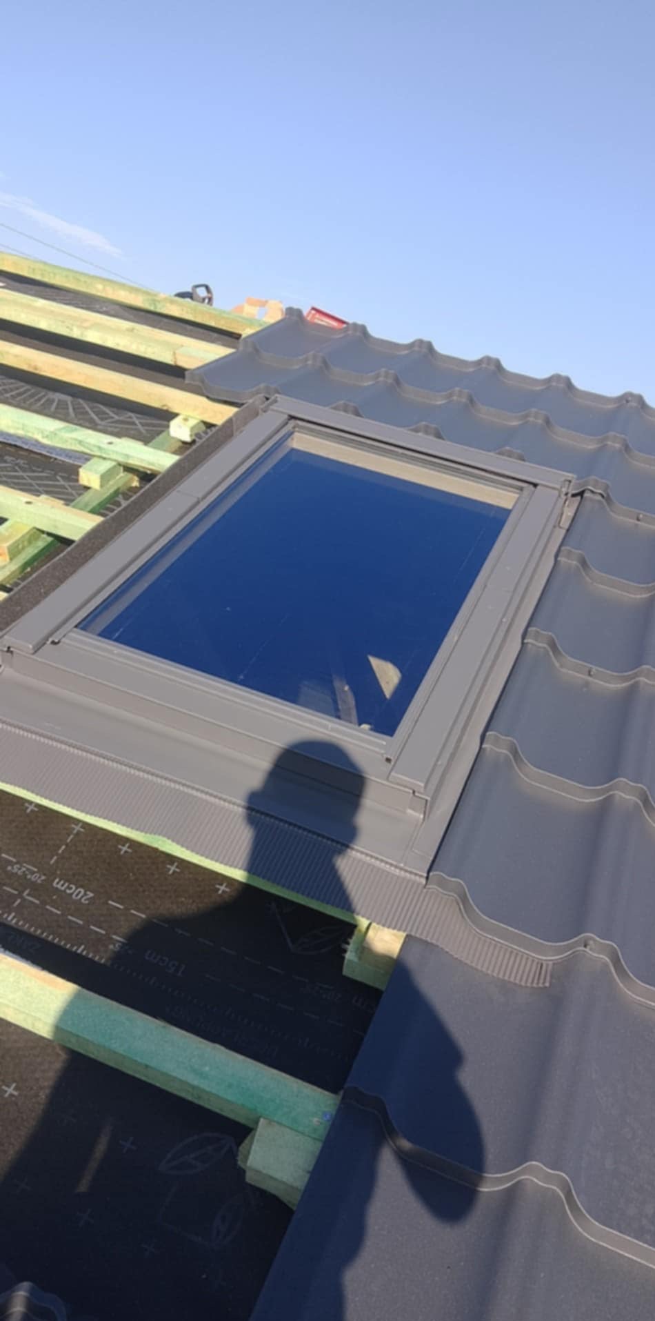 Montaż okna dachowego na nowym dachu z ciemnej blachy falistej, widoczna więźba i membrana dachowa, cień osoby fotografującej na dachu, błękitne niebo.