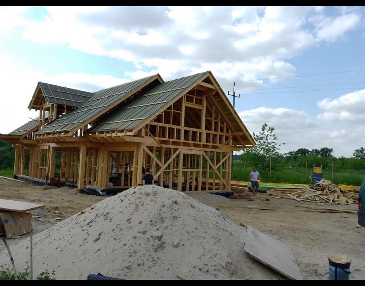 Wznoszenie drewnianej konstrukcji domu jednorodzinnego z częściowo ułożonym pokryciem dachowym z dachówki ceramicznej, widoczny szkielet więźby dachowej i ściany, na pierwszym planie pryzma piasku...