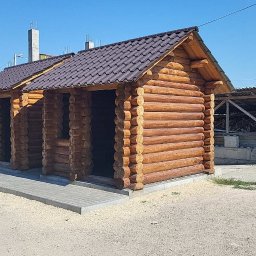 OLEKSANDR SMERECHUK PraceBud - Dwa połączone budynki z bali z brązowym dachem, widoczne na tle placu budowy i mężczyzny pracującego przy układaniu kostki brukowej.