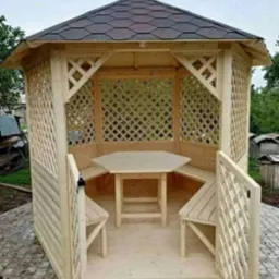 Drewniana altana ogrodowa z heksagonalnym stołem i ławkami, zadaszona gontem bitumicznym, widok z zewnątrz, otwarte wejście. Elementy kratowe i boazeria.
