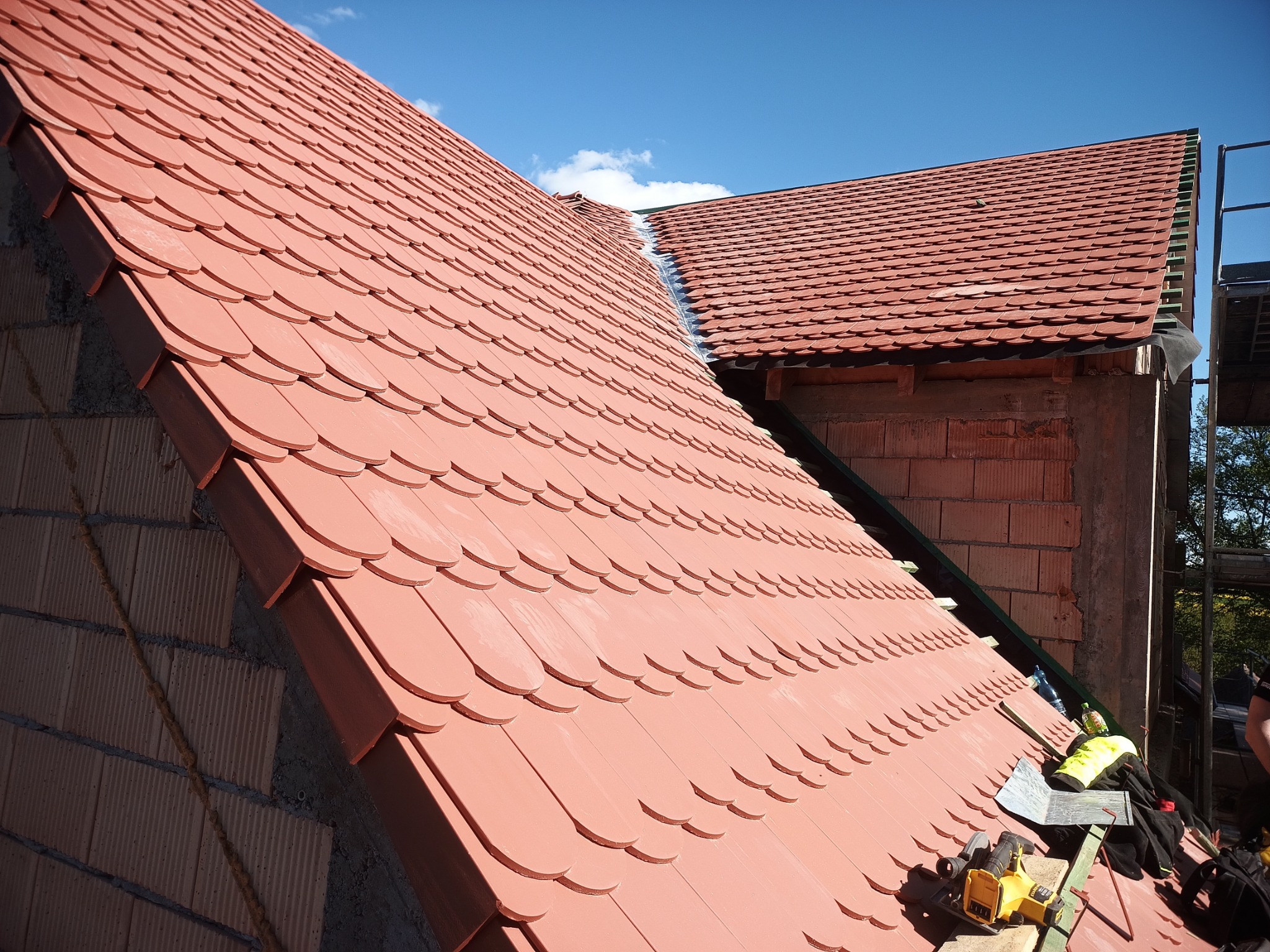 Nowy dach z czerwonej dachówki karpiówki w trakcie montażu. Widoczne narzędzia i fragment rusztowania przy budynku z cegły. Błękitne niebo w tle.