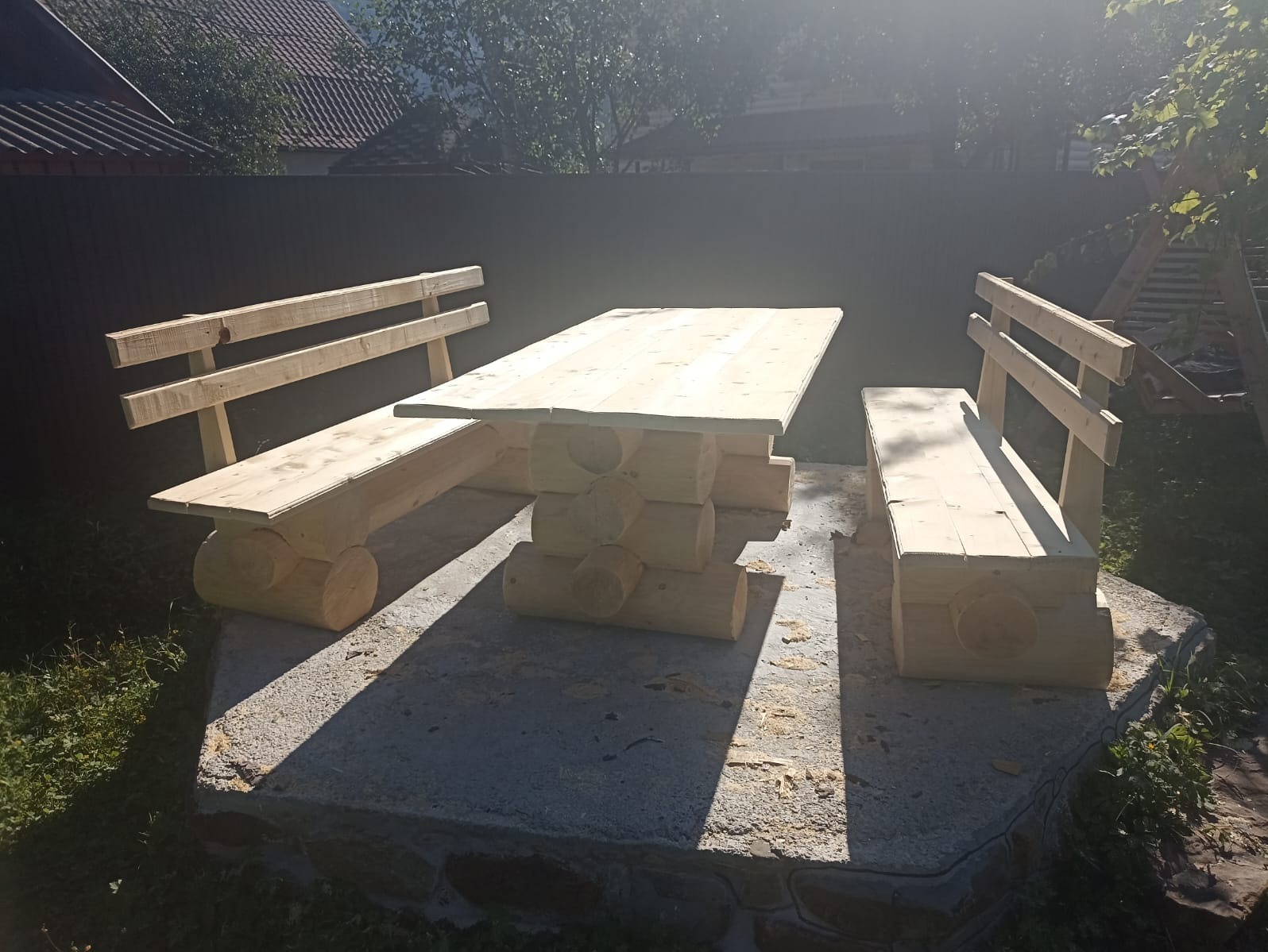 Drewniany stół z ławkami, wykonany z bali, ustawiony na betonowym podwyższeniu, w ogrodzie. Widoczne słońce i cień, co dodaje naturalnego charakteru.