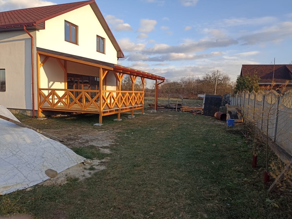 Biały dom z drewnianą werandą w stylu rustykalnym, widok z ogrodu. Elementy drewniane pomalowane na ciepły, miodowy kolor. Tło: błękitne niebo z chmurami.