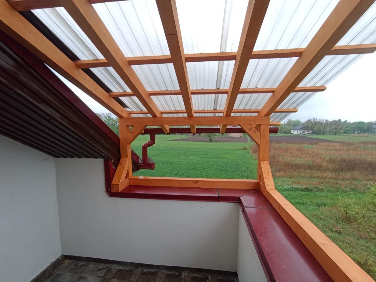 Zadaszenie balkonu z drewnianą konstrukcją i przezroczystym pokryciem, widok na zielone pole. Elementy brązowej blachy dachowej i rynny.
