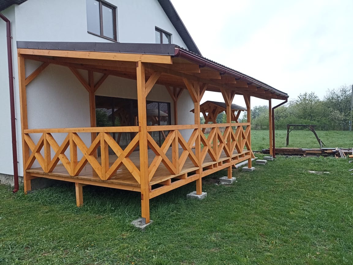 Drewniana weranda z balustradą w kształcie krzyży przy białym domu. Widoczne brązowe rynny i elementy dachu. Na trawniku leżą deski.