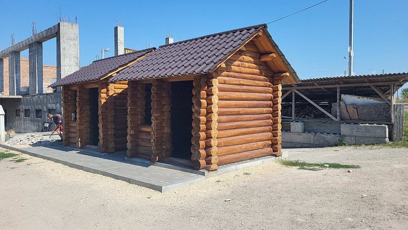 Dwa połączone budynki z bali z brązowym dachem, widoczne na tle placu budowy i mężczyzny pracującego przy układaniu kostki brukowej.