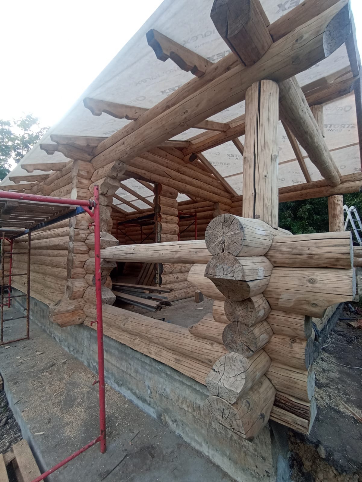 Surowa konstrukcja domu z bali drewnianych w trakcie budowy, widoczne rusztowania i elementy więźby dachowej zabezpieczone folią. Detal łączenia bali.