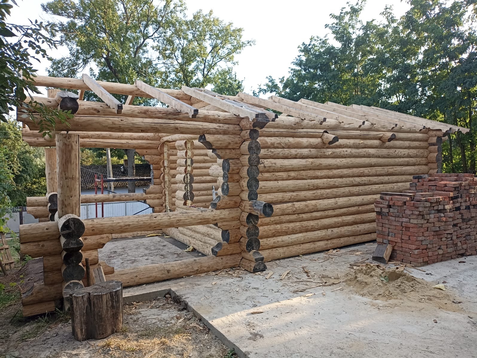 Drewniana konstrukcja domu z bali w trakcie budowy, widoczne elementy więźby dachowej i stos cegieł obok budynku. Całość w naturalnym otoczeniu drzew.