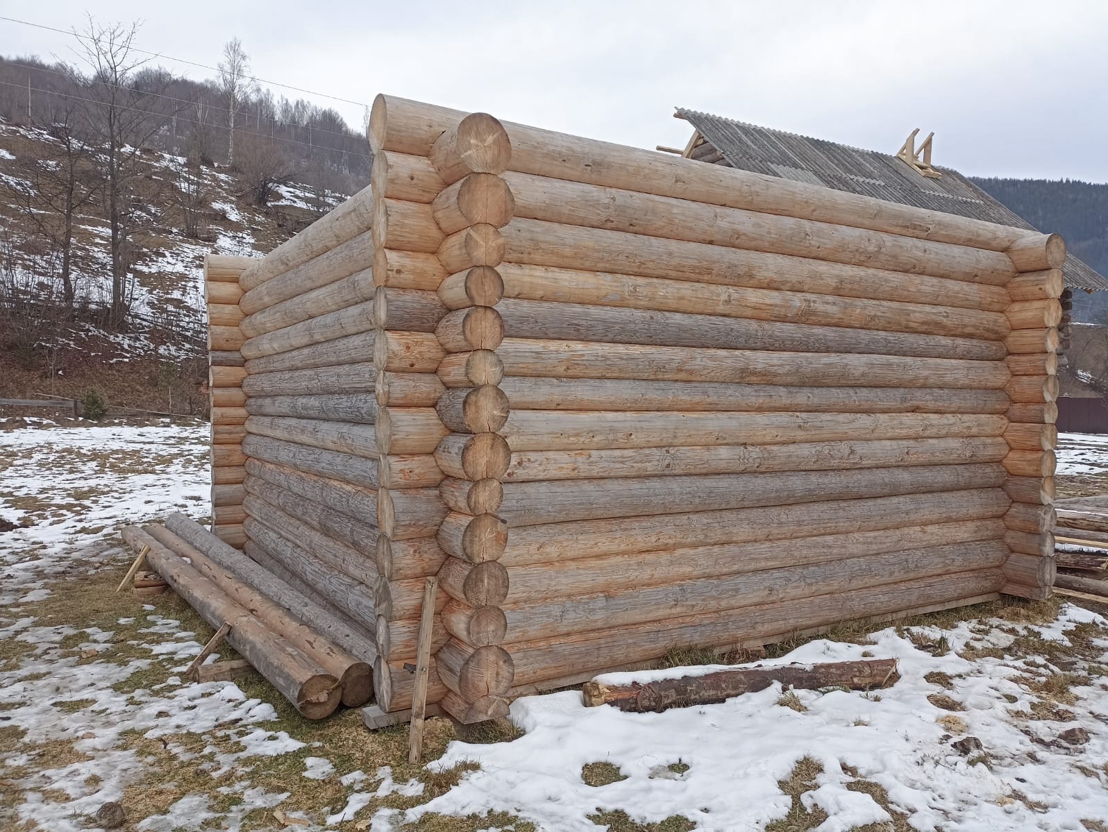 Surowy stan budowy domu z bali, widoczne połączenia bali na węgłach, częściowo ośnieżony teren, w tle górzysty krajobraz i fragment dachu.