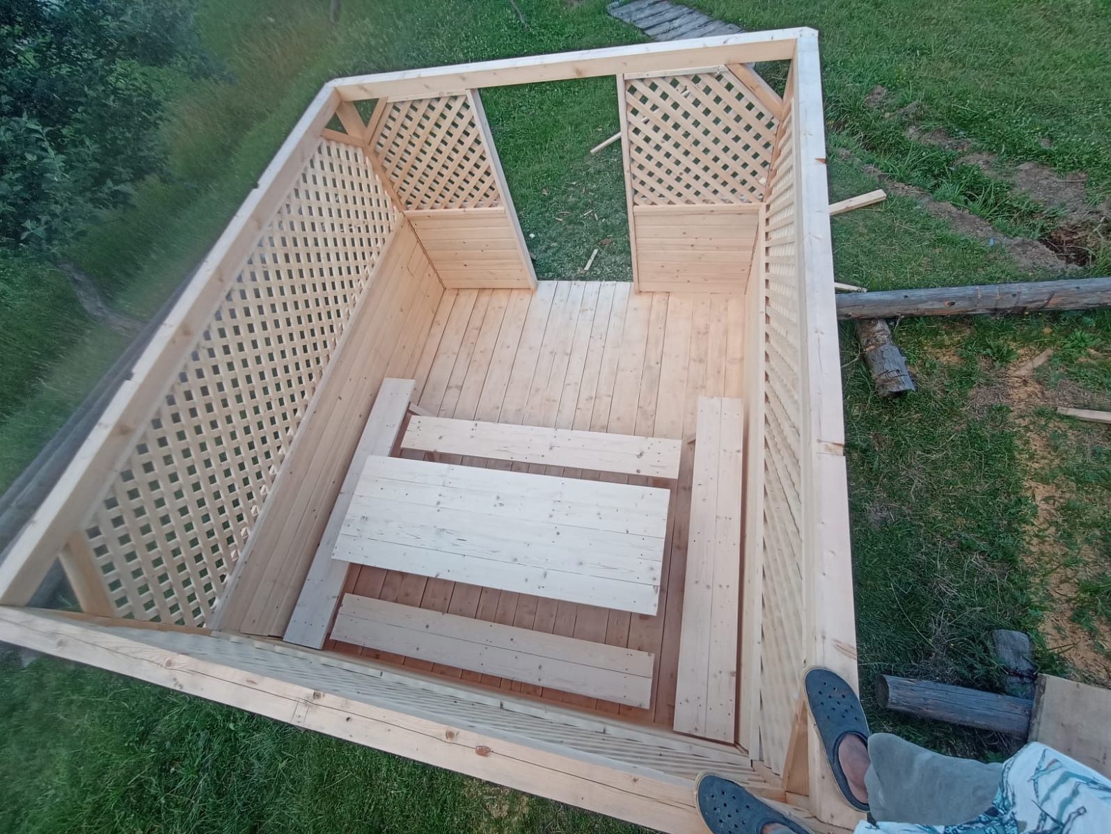 Wysoka perspektywa na drewnianą altanę z kratownicą, częściowo zmontowaną, z widocznymi deskami podłogowymi i elementami konstrukcyjnymi. Podłoże trawiaste.