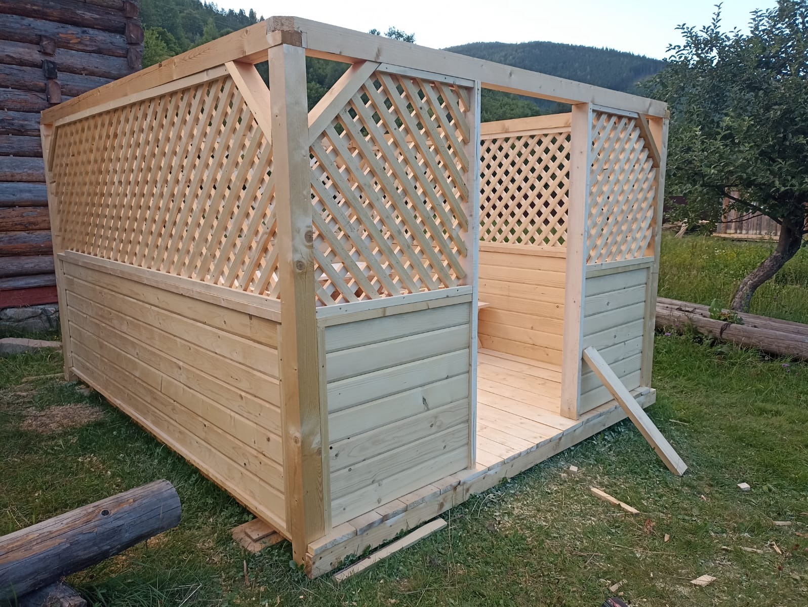 Drewniana altana w trakcie montażu, z widoczną konstrukcją szkieletową i kratownicą, na tle zieleni. Elementy z jasnego drewna, surowy stan wykończenia.