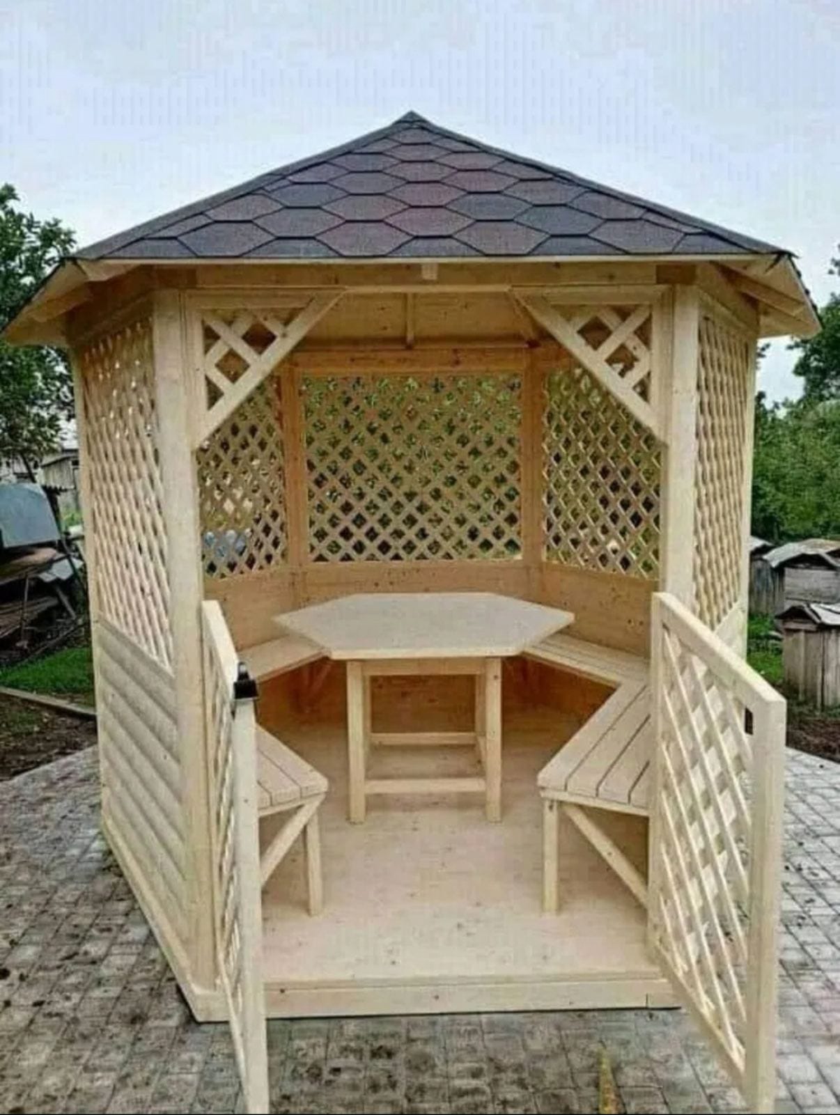Drewniana altana ogrodowa z heksagonalnym stołem i ławkami, zadaszona gontem bitumicznym, widok z zewnątrz, otwarte wejście. Elementy kratowe i boazeria.