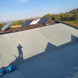 Artur Górak Backup-Dach - Płaski dach pokryty papą z trzema panelami słonecznymi, widoczne worki z odpadami i cień osoby robiącej zdjęcie, w tle maszt telekomunikacyjny i zalesione wzgórze.